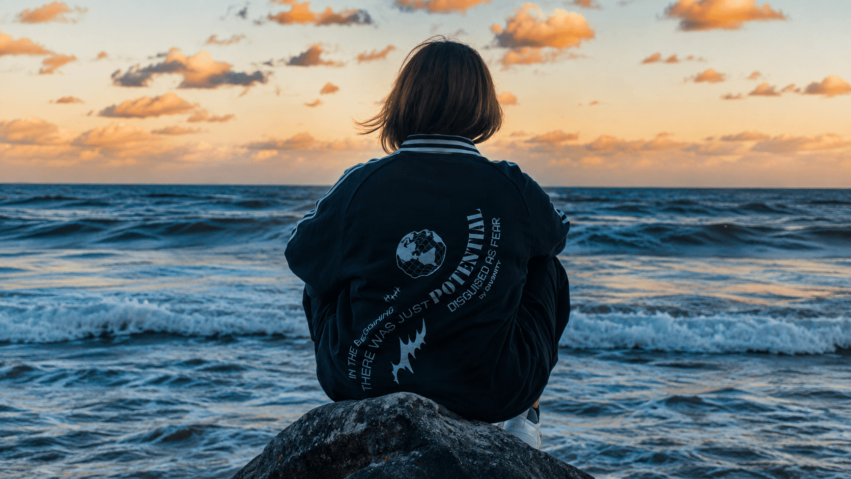 Mayte sitting on a rock facing the ocean at sunset, wearing a DIV3NITY jersey